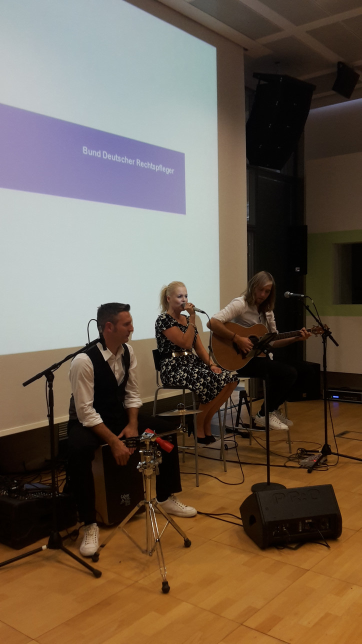 Drei Musiker (Percussion, Gesang, Gitarre) performen in einem Raum vor Publikum